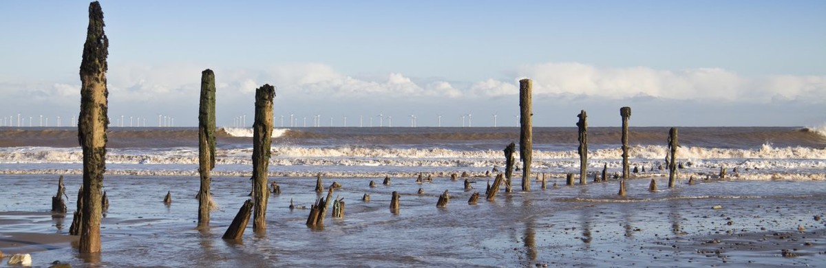 Remnants of a pier sticking out of the sea shore with wind turbines in the distance