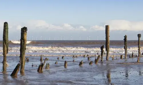 Remnants of a pier sticking out of the sea shore with wind turbines in the distance