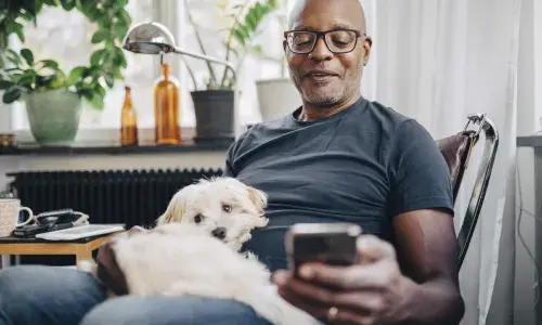 Smiling retired senior male using smart phone while sitting with dog in room at home