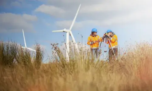 Man and woman standing in grassy field in front of windfarms, wearing hard hats and high-vis jackets with a survey camera - used for the National Grid story 'Now is the time to recruit a Net Zero Energy Workforce to power a greener future'