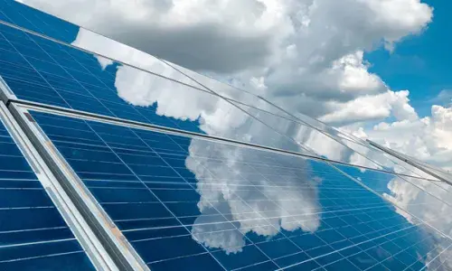 Solar panels beneath a blue, cloudy sky with the clouds reflecting in the panels