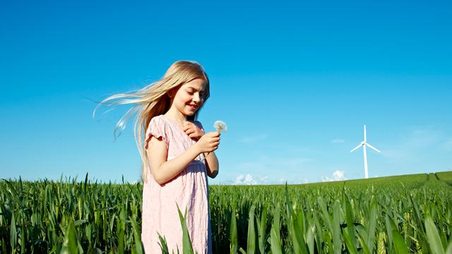 EnergyExplained_WhyNetZeroImportant_640x360.jpg Girl holding a dandelion clock standing in a field with wind turbine in the background
